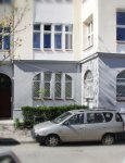 Widok na wejście do kamienicy, w której znajduje się dzienny dom pomocy. Szaro-kremowa fasada z dwoma rzędami dużych okien. Brama wejściowa ze sklepieniem łukowym. Przed budynkiem rząd zaparkowanych samochodów